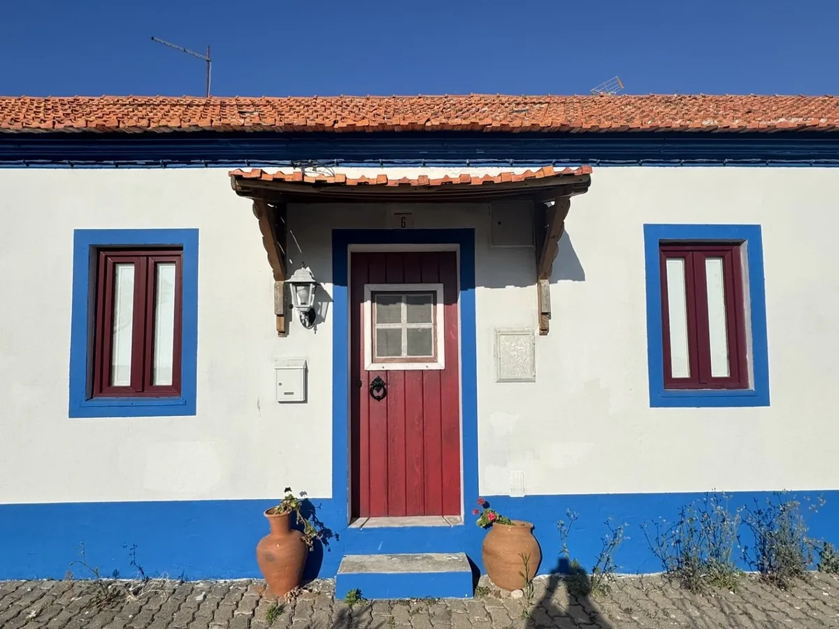 A white-and-blue Comporta house with a red door, terracotta pots in the warm afternoon light
